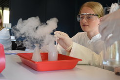 Scientific experiment with smoke development in a beaker, held by a pupil in laboratory clothing and protective gloves.