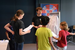 In one room, five people, four students and one supervisor, are engaged in a discussion. In the background, a row of white lab coats hangs on a clothes rail in front of a colorful poster on the wall.