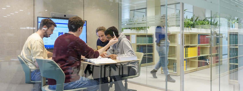Postdocs Four people sit in a modern room with glass walls, surrounded by bookshelves, and work together. Behind them are bookshelves and a person walking past. A screen in the room shows additional data.